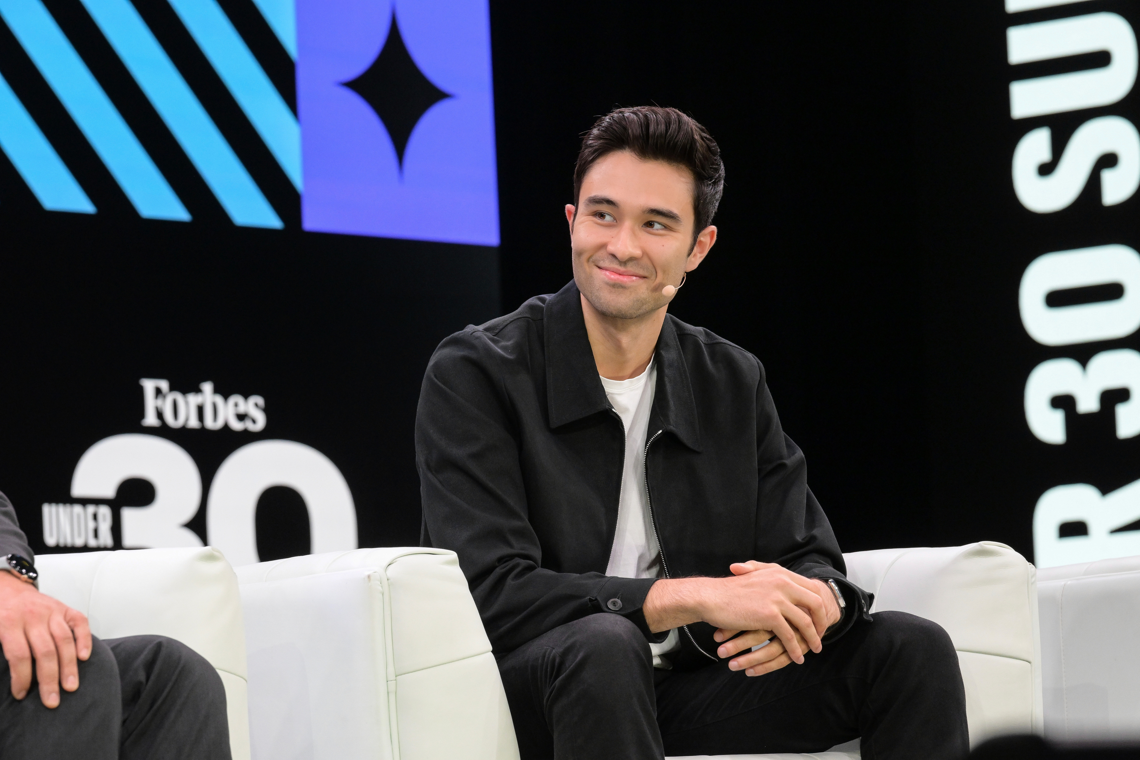 Rowan Cheung seated onstage at a Forbes event, wearing a headset microphone