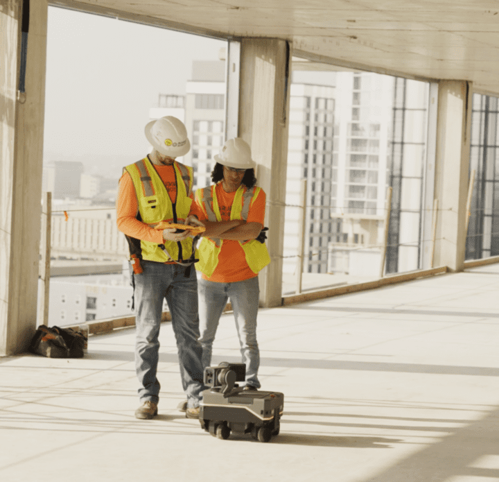 Two hard-hatted field workers reviewing a tablet beside a Rugged layout robot