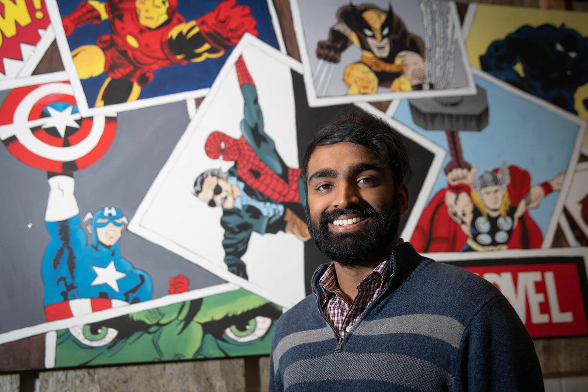 Rupesh Jeyaram smiling in front of comic-book artwork at Caltech