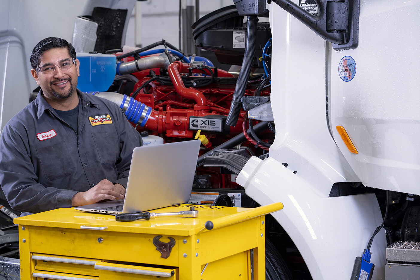 Rush Truck Centers technician Adam beside an open truck engine, using a laptop on a yellow tool cart