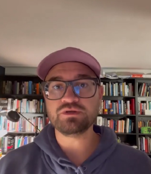 Ryan David Mullins wearing glasses and a cap, seated in front of bookshelves