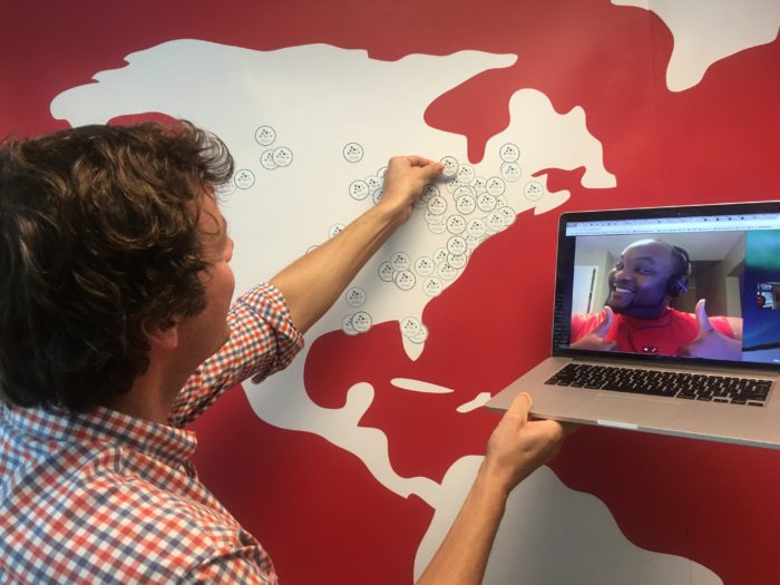Ryan Jones at a wall map with hand-drawn faces, holding a laptop showing a colleague on a video call