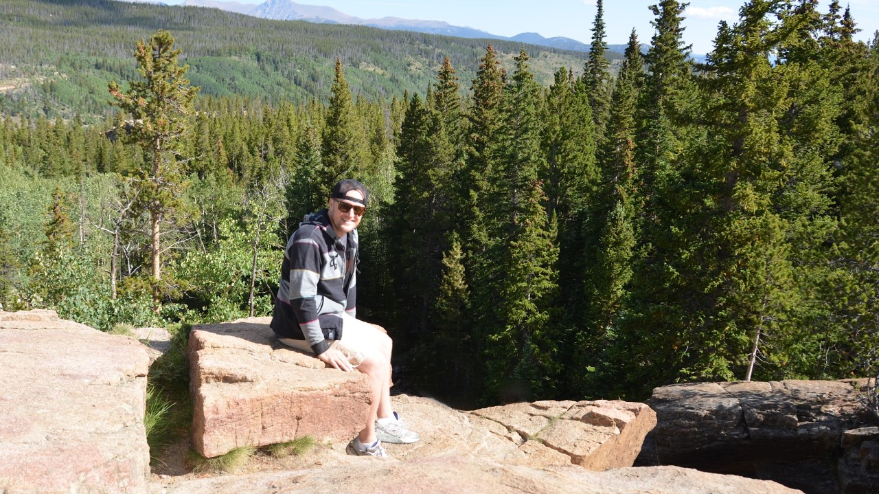 Ryan Medine seated on a rocky overlook above a forested mountain landscape