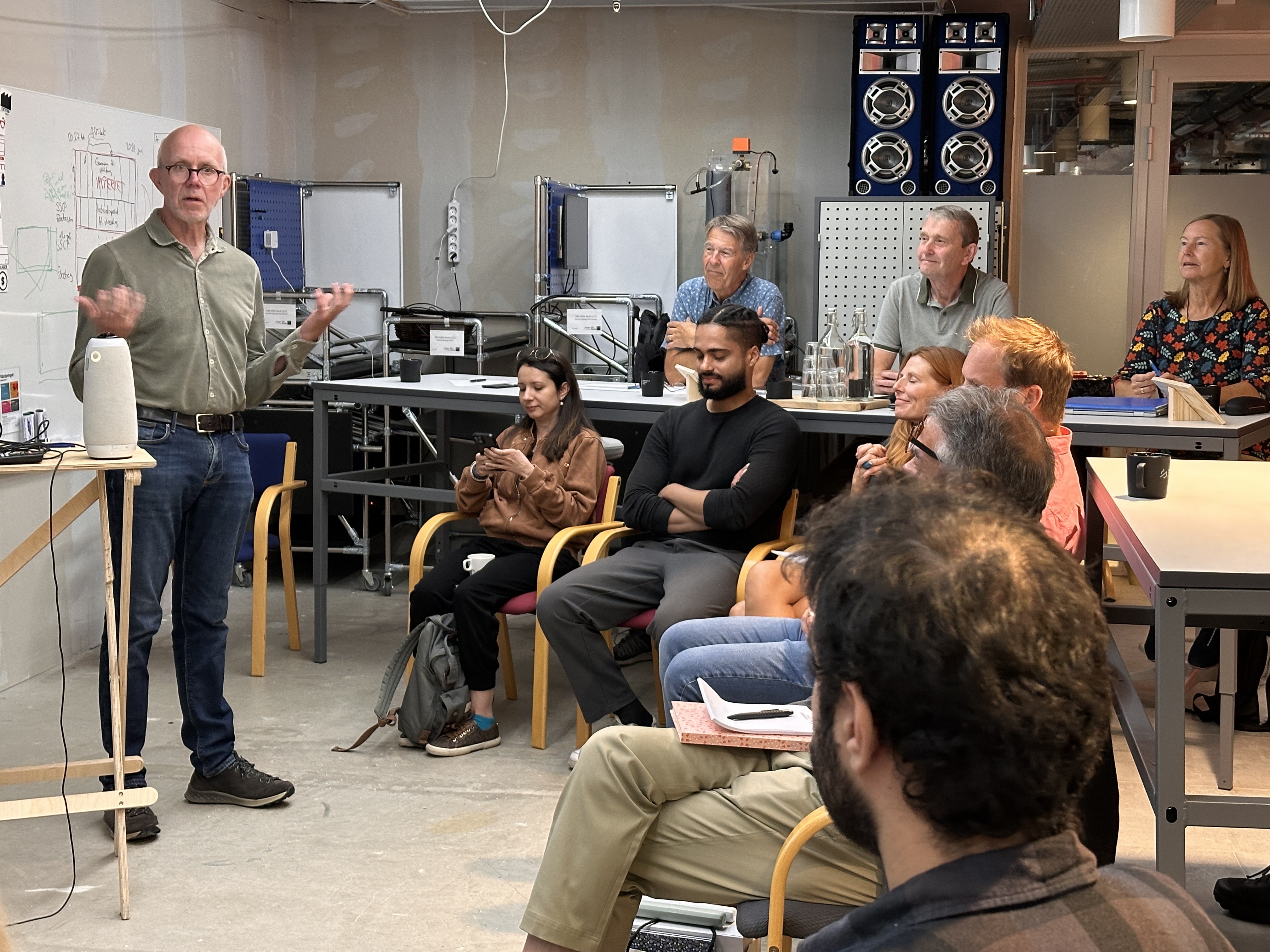 A presenter speaks with startup participants in a workshop with equipment and a whiteboard