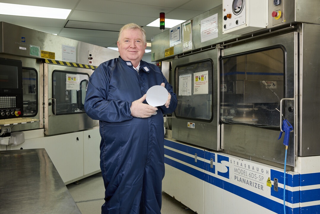 Sam Anderson in a blue cleanroom suit holding a silicon wafer inside the IceMOS manufacturing facility