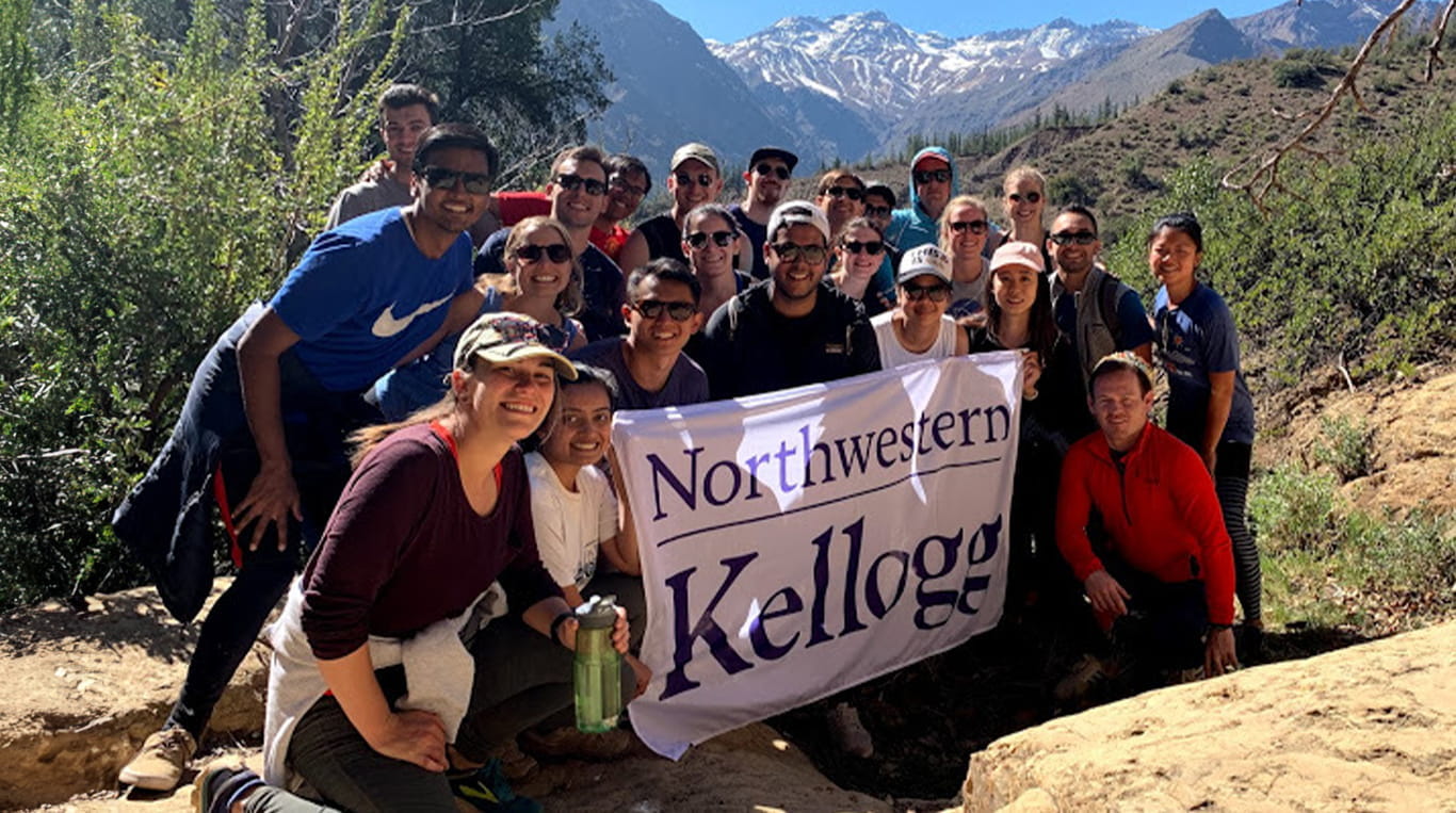 Kellogg students standing on a rocky hike with a purple school flag