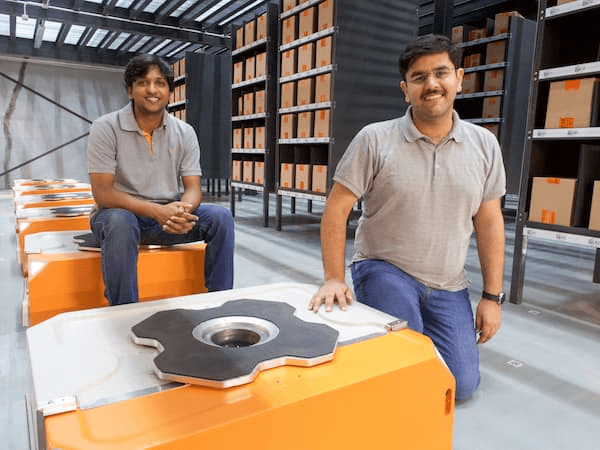 Akash Gupta seated on a warehouse robot beside Samay Kohli standing among inventory shelves