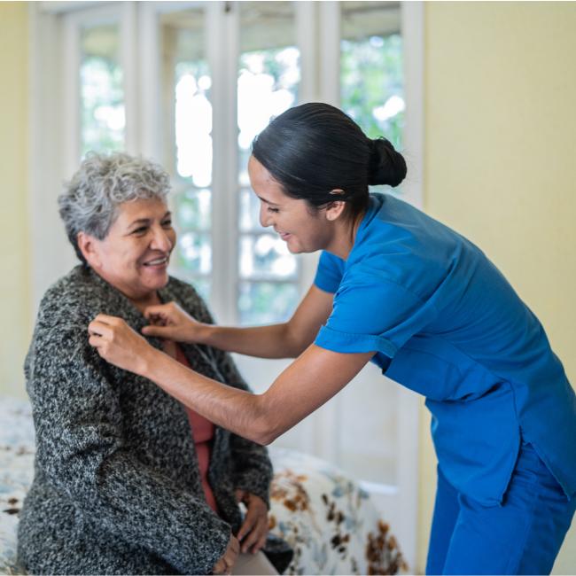 A caregiver helping an older woman fasten her cardigan in a home