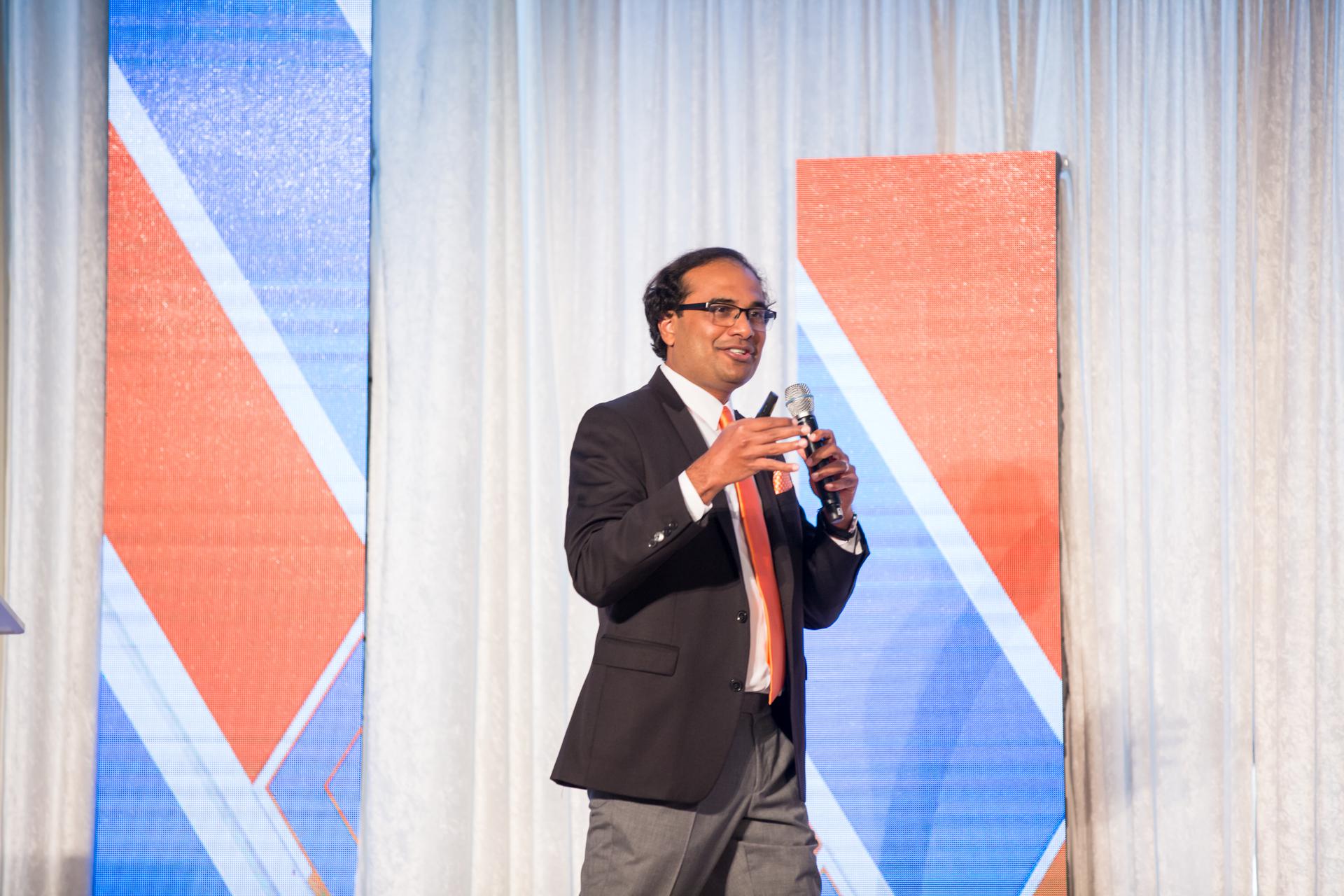 Sashi Narahari speaking into a microphone on a stage with blue and orange geometric panels