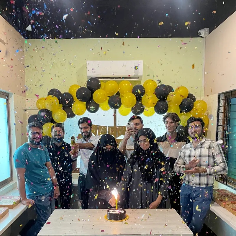 Sastaticket team members celebrating around a cake under yellow and black balloons and falling confetti