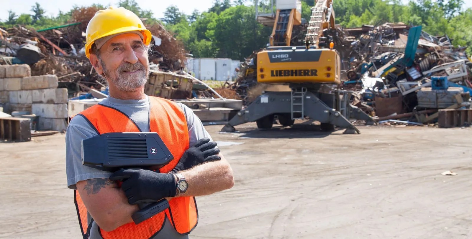 A recycling-yard worker holding a SciAps handheld analyzer among piles of scrap metal