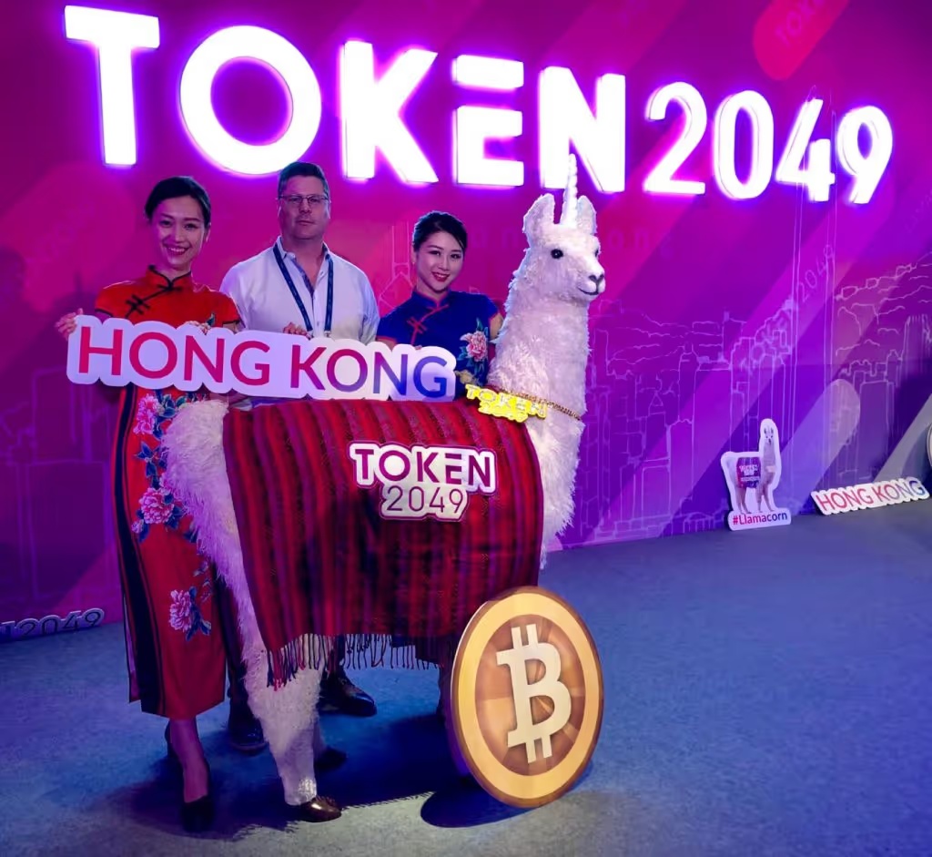 Scott Bambacigno at TOKEN2049 in Hong Kong beside event hosts and a llama display