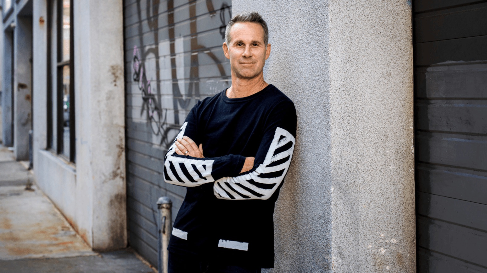 Scott Cutler in a black top with white sleeve stripes, leaning beside a graffiti-marked doorway