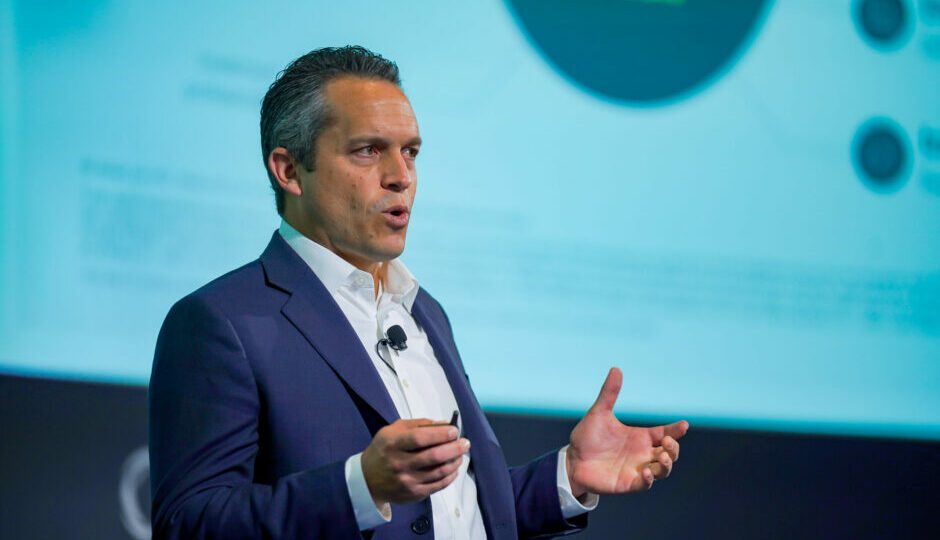 Scott Hart speaking onstage, holding a presentation remote and gesturing with his other hand