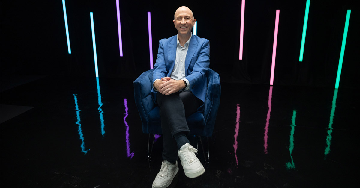 Scott Russell seated on a blue chair in a studio with colored vertical lights