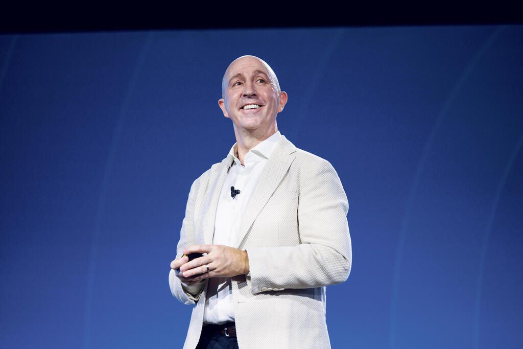 Scott Russell speaking onstage, holding a presentation remote against a blue backdrop