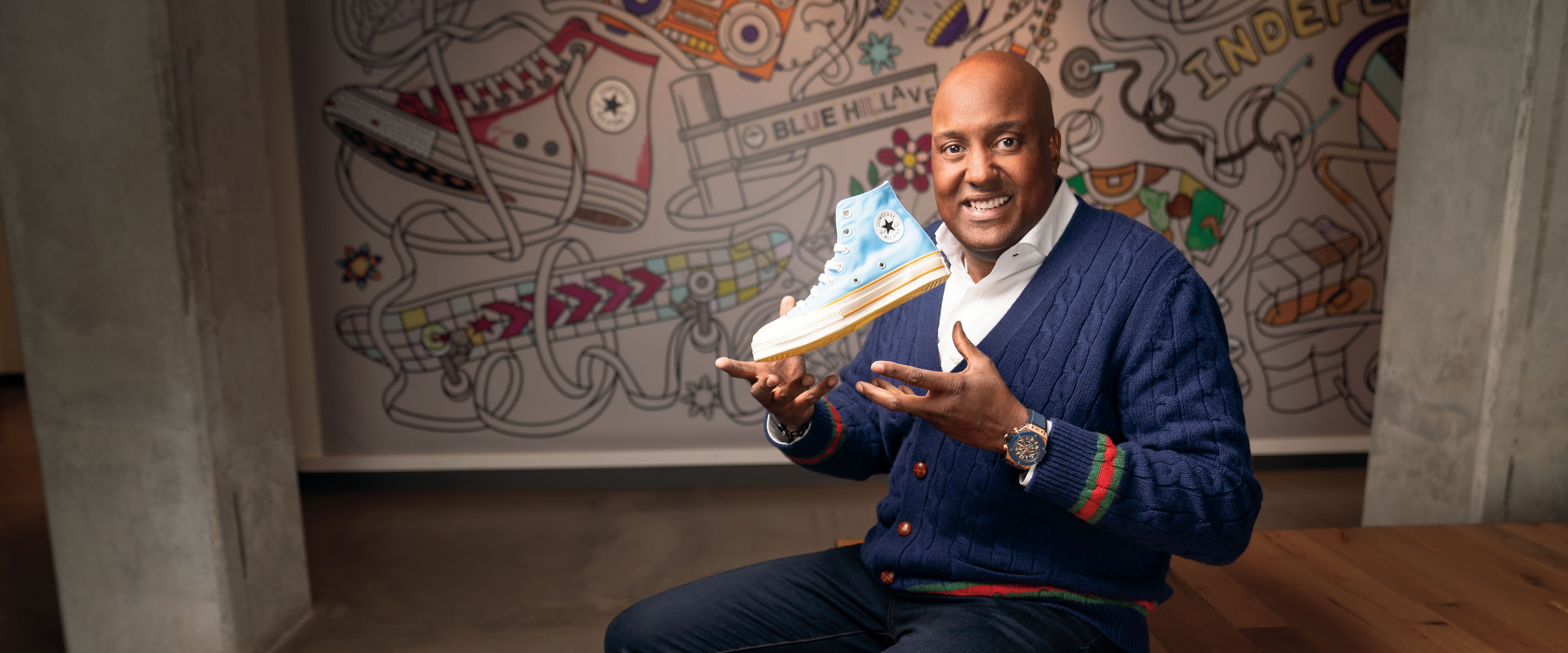 Scott Uzzell holding a blue Converse high-top in front of a wall of illustrated sneakers
