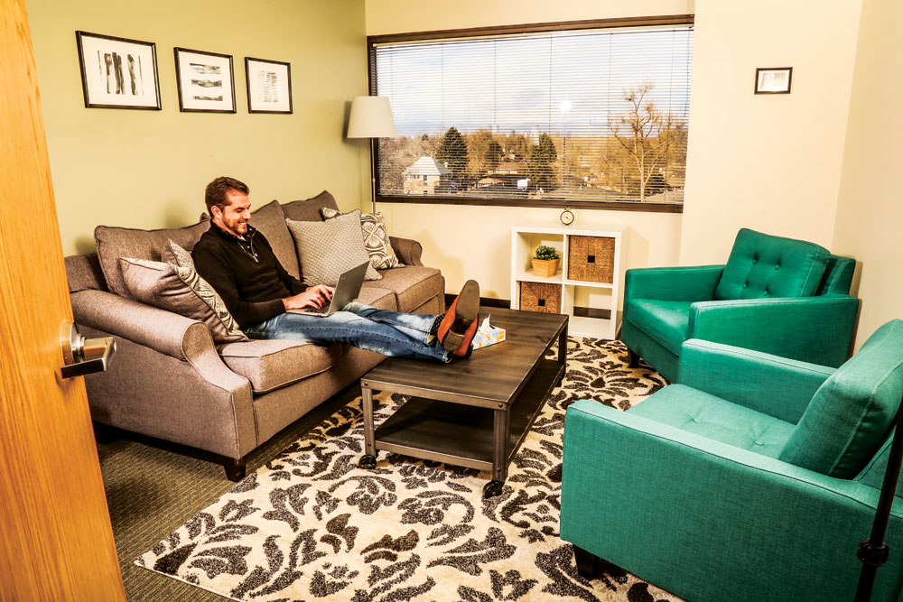 Sean Boyd uses a laptop while seated on a gray sofa, with his feet resting on a coffee table