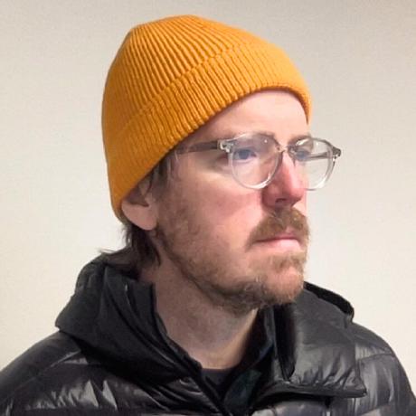 Sean Conaty wearing glasses and a mustard-colored knit hat in his GitHub profile photograph