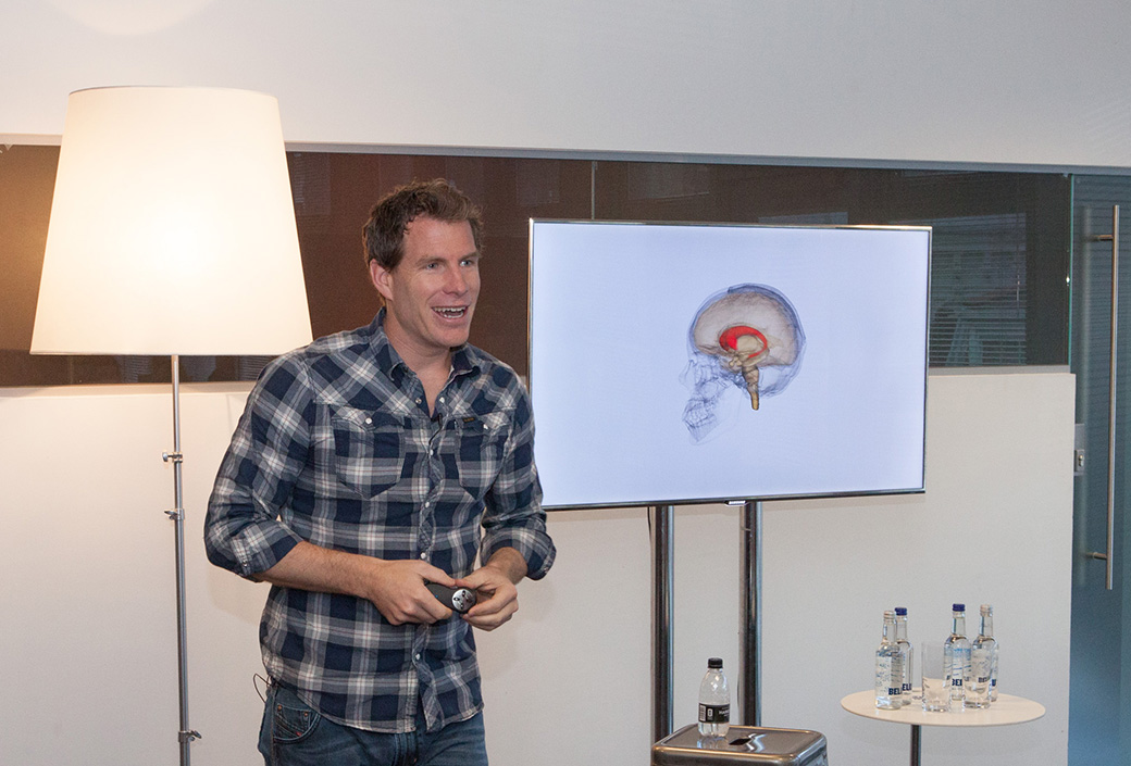 Sean Gourley speaking beside a screen showing a brain diagram at an AKQA event