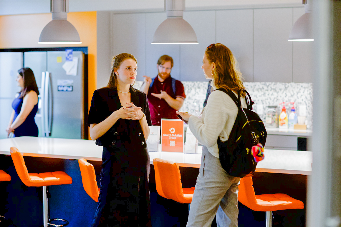 Two attendees talking beside orange stools at a Search Solution Group gathering