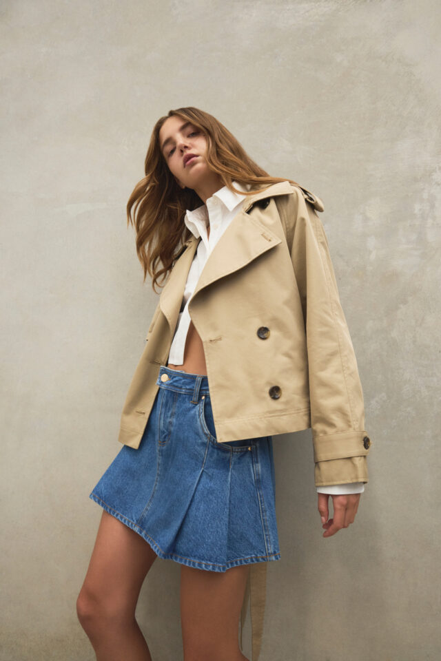 Aje fashion product photograph showing a model in a cropped beige trench jacket and denim skirt