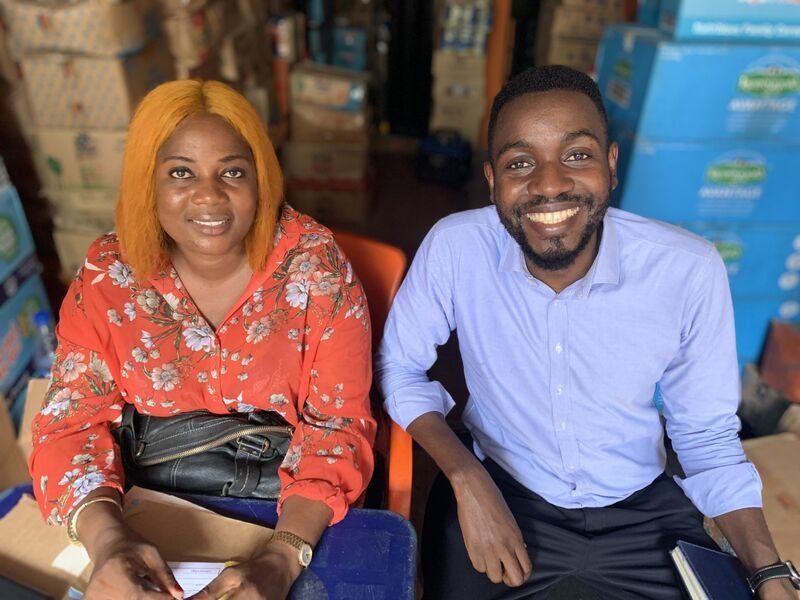 Sefa Ikyaator, seated on the right beside a woman in a shop surrounded by boxes of stock.