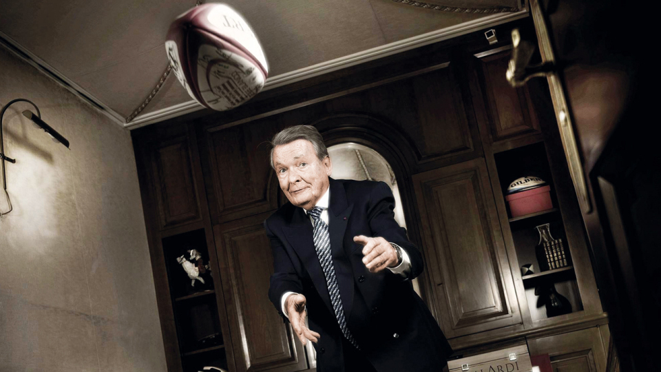 Serge Kampf in a suit tossing a rugby ball in a wood-paneled room