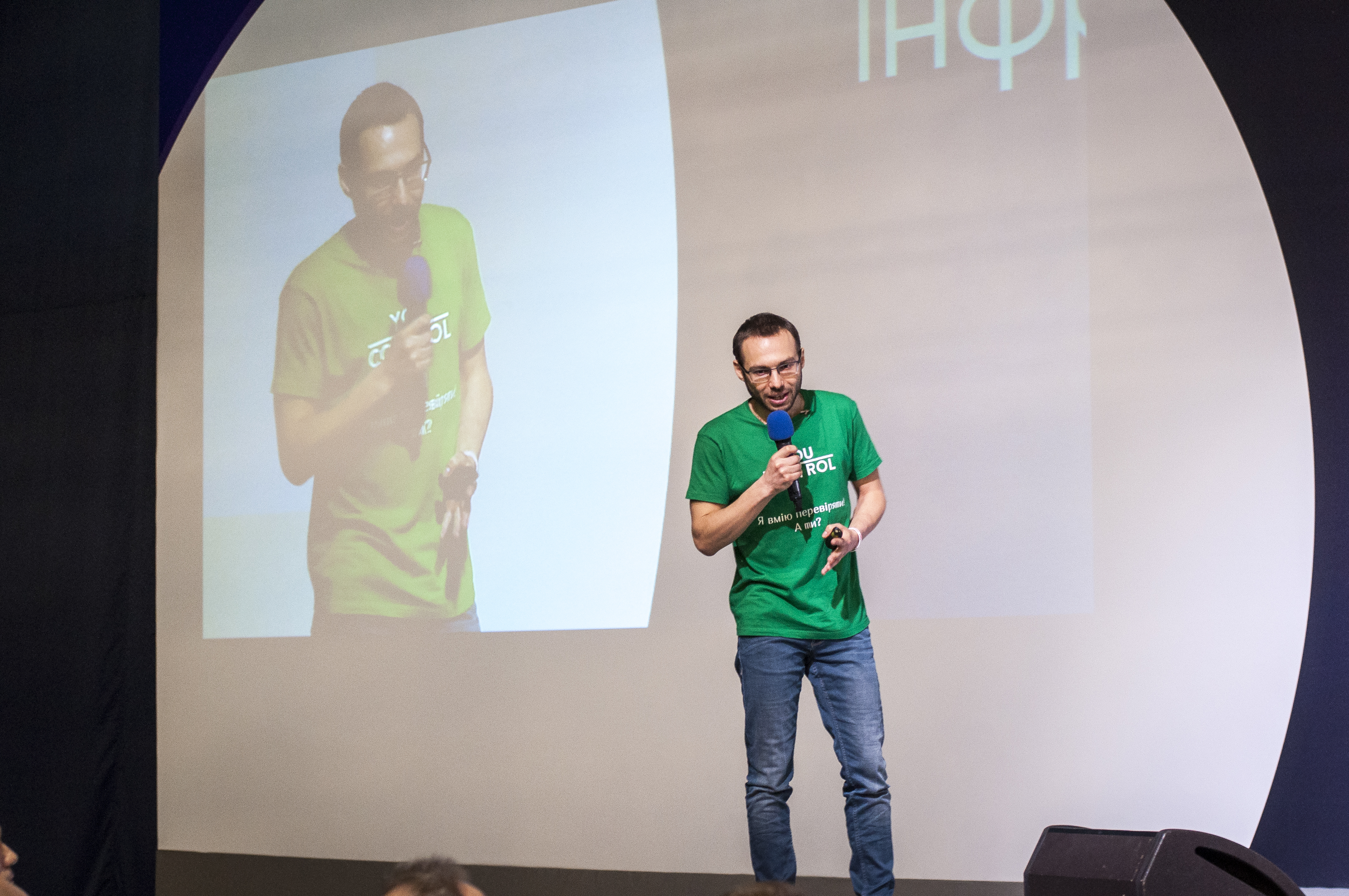 Sergiy Milman speaking on stage in a green YouControl T-shirt, with his image projected behind him