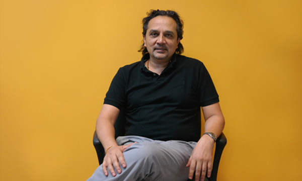 Shailesh Naik seated against a yellow wall, wearing a black polo shirt