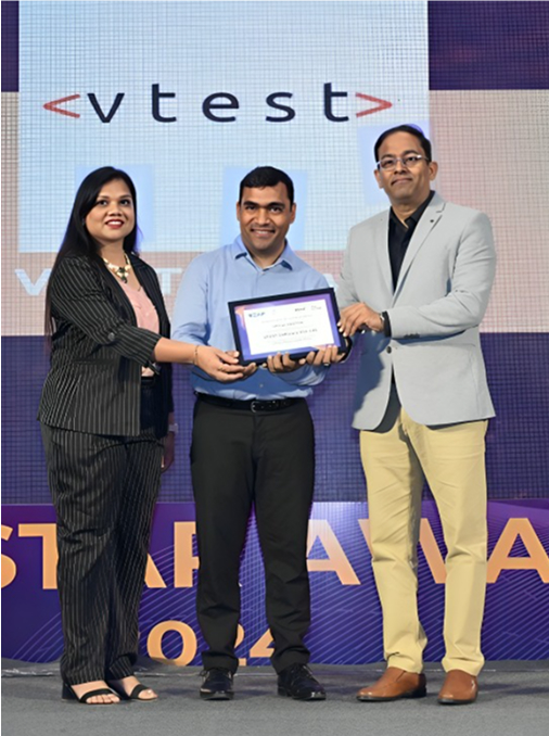 Shakil Hanjgikar, centre, holding a framed certificate with two people on a VTEST-branded stage