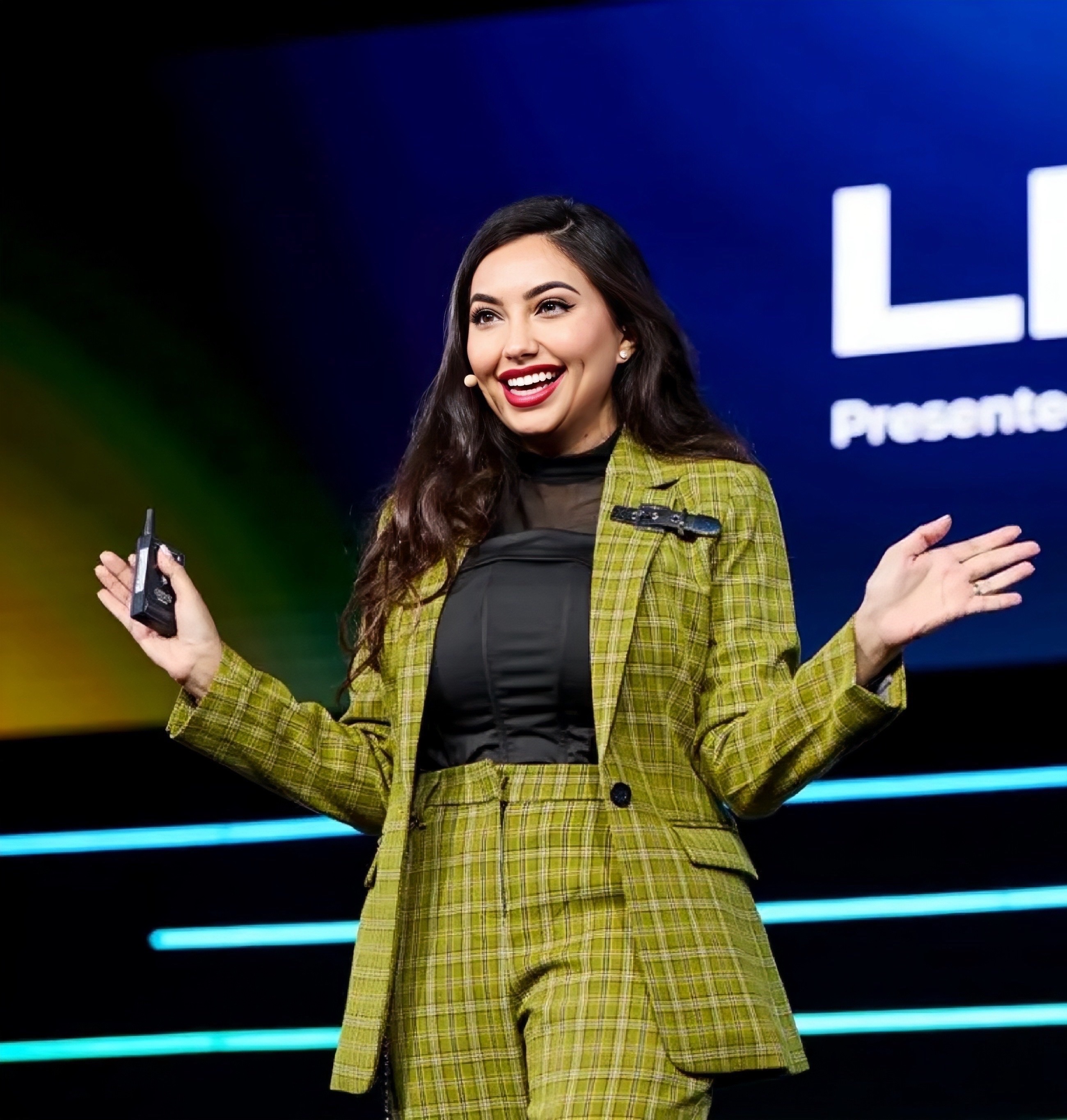Shama Hyder speaking on stage in a green checked suit, holding a presentation remote