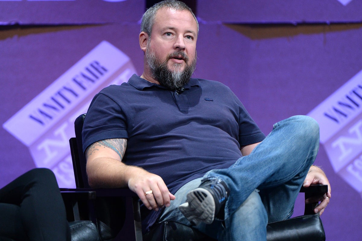 Shane Smith seated onstage during a media conference, speaking before a purple backdrop