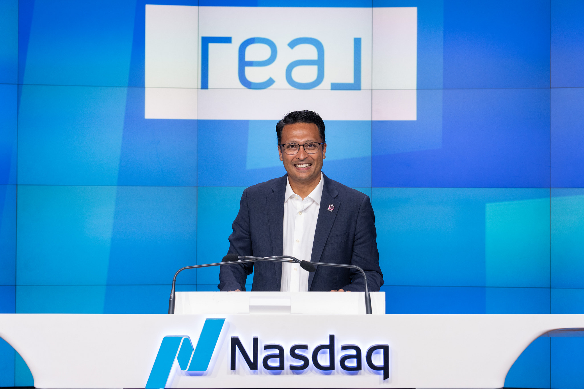 Sharran Srivatsaa speaking at a Nasdaq podium in front of a Real display