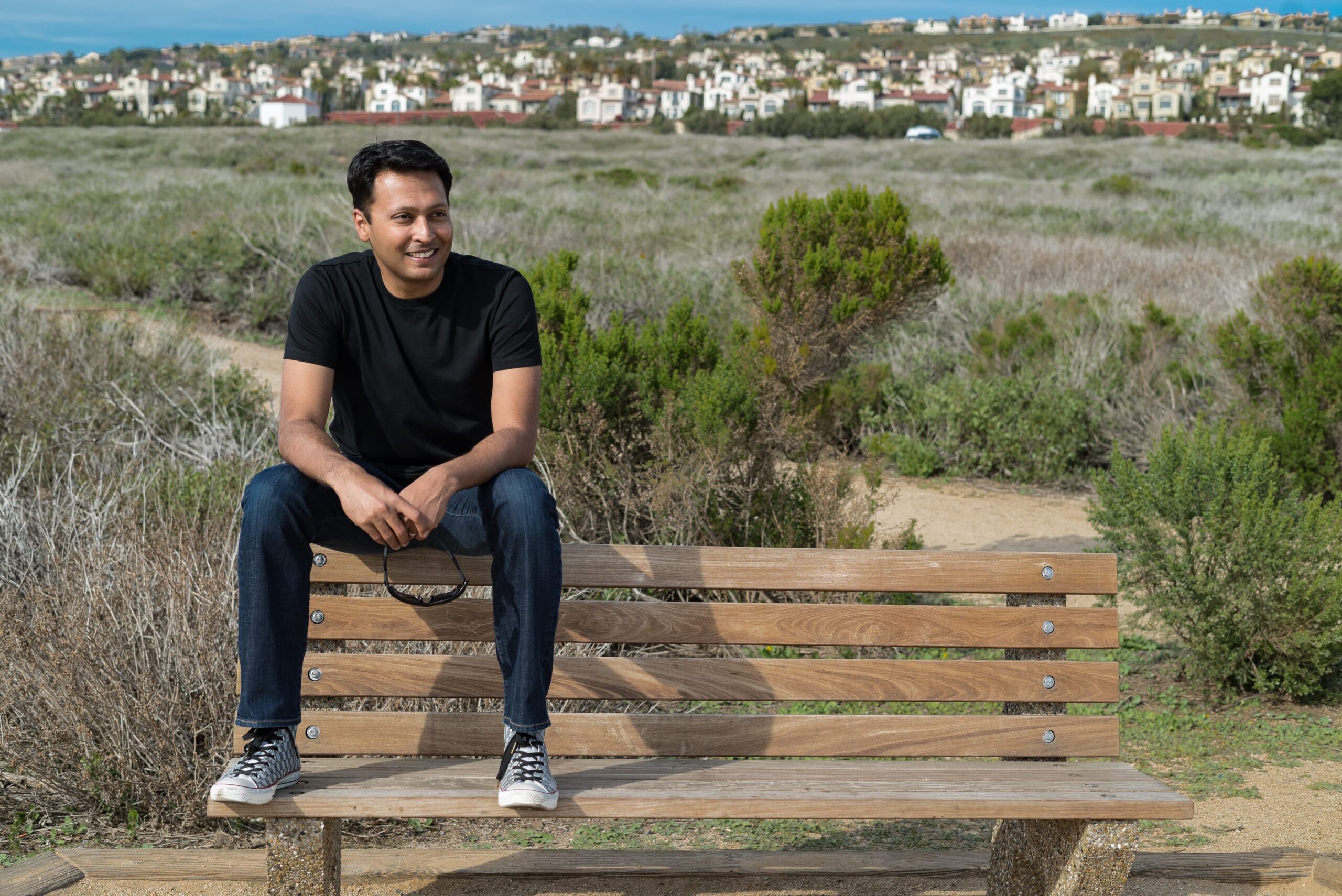 Sharran Srivatsaa seated on a wooden bench with coastal houses in the distance