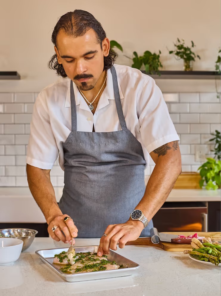 A cook in a gray apron brushing herbs onto fish fillets in a kitchen.