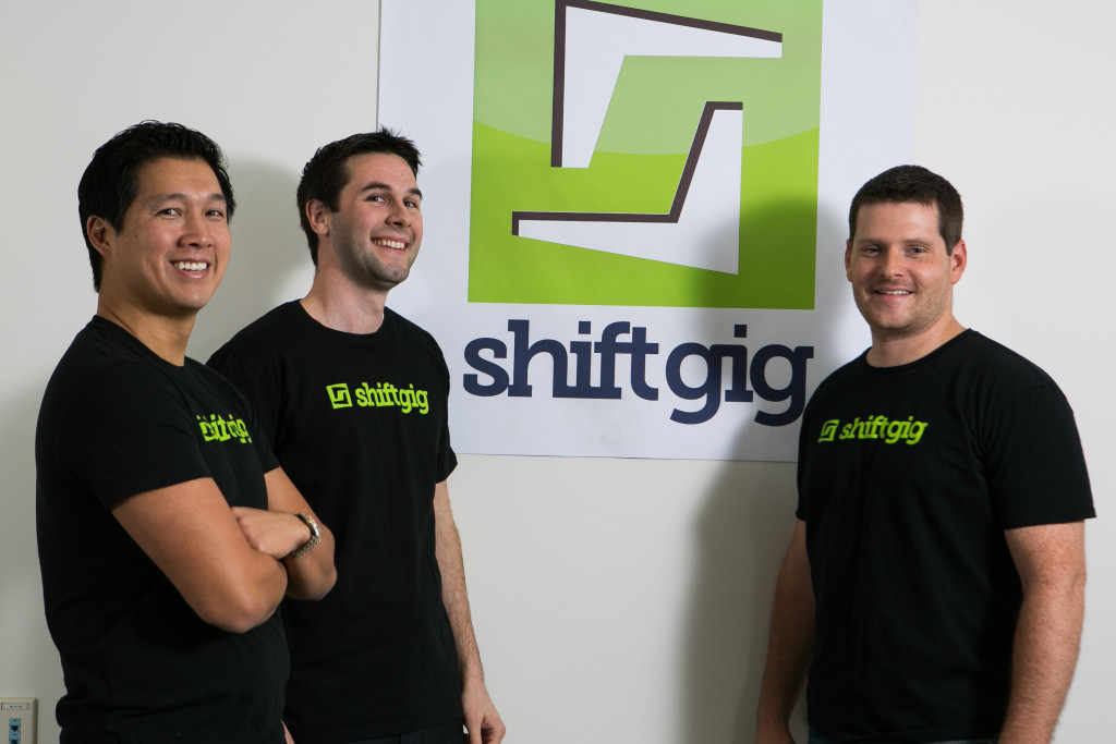 Three members of Shiftgig’s early founding team in matching black company T-shirts beside its original logo.