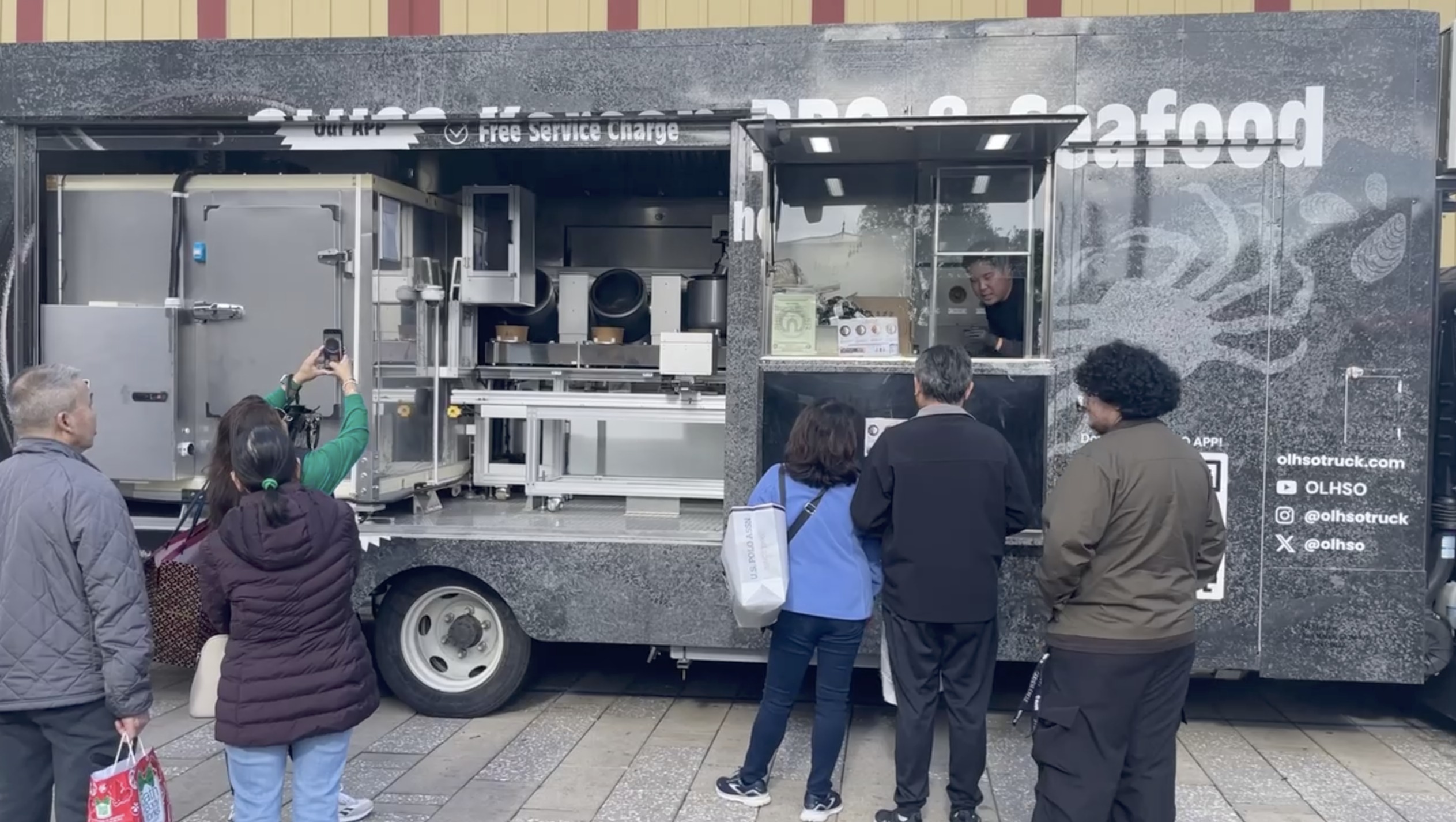 Customers gather beside an open OLHSO automated food truck, with its robotic cooking drums visible inside