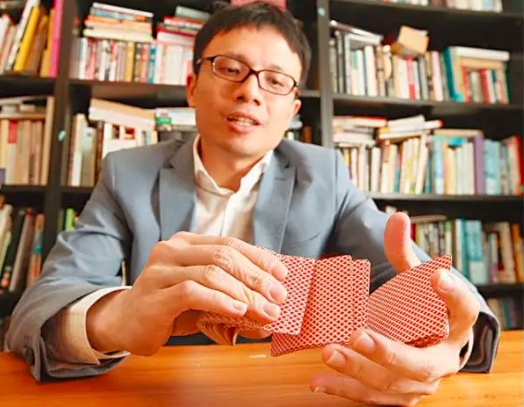 Shing Chow holding red-backed playing cards at a table in front of bookshelves