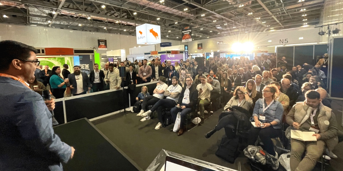 Sean Mallapurkar speaking into a microphone before a seated audience at an industry exhibition