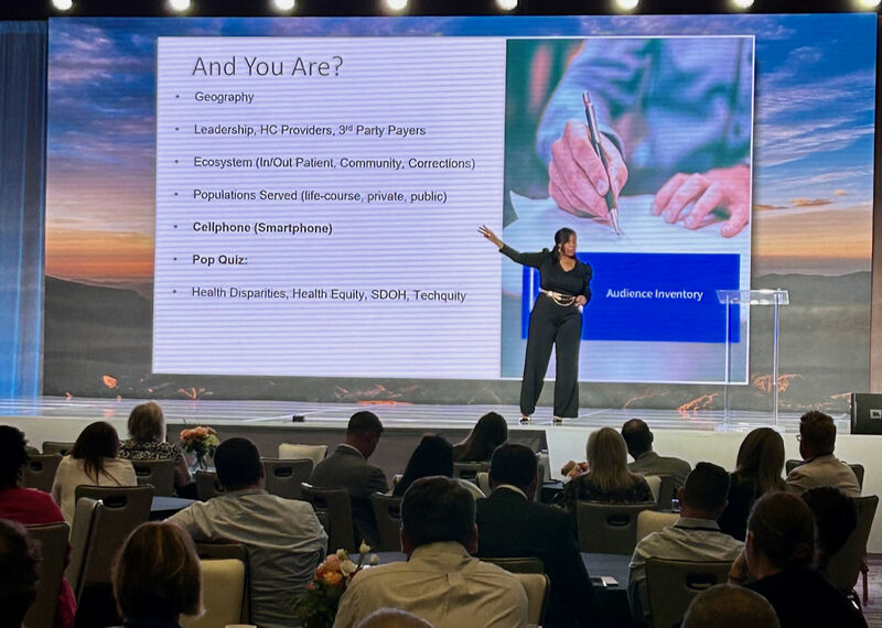 A speaker on the WellSky CareForum 2023 stage addresses an audience seated at round tables
