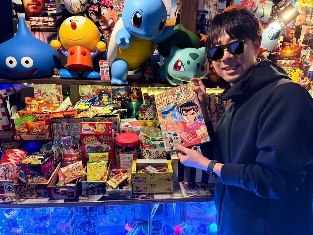 Shunsuke Oyu holding a manga magazine among character figures and memorabilia at Showa Bar in Okinawa