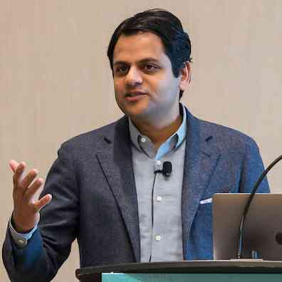 Shyam Rao speaking behind a lectern, gesturing beside a laptop