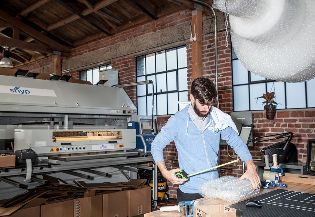 A Shyp worker measuring a wrapped item beside a box-making machine at the company’s packing facility