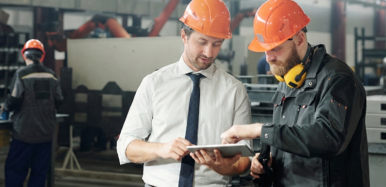 Two industrial workers consult a tablet beside factory equipment