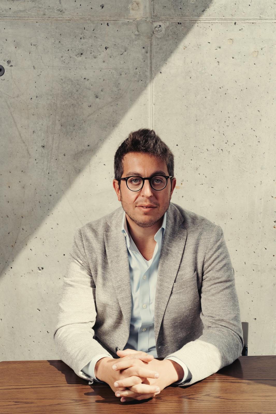 Simcha Hyman seated at a wooden table, wearing glasses and a gray jacket