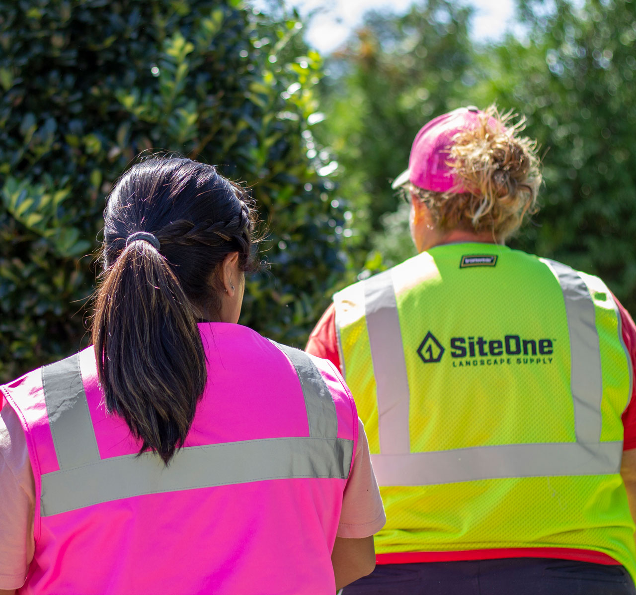 Two SiteOne associates wearing pink and yellow safety vests among greenery