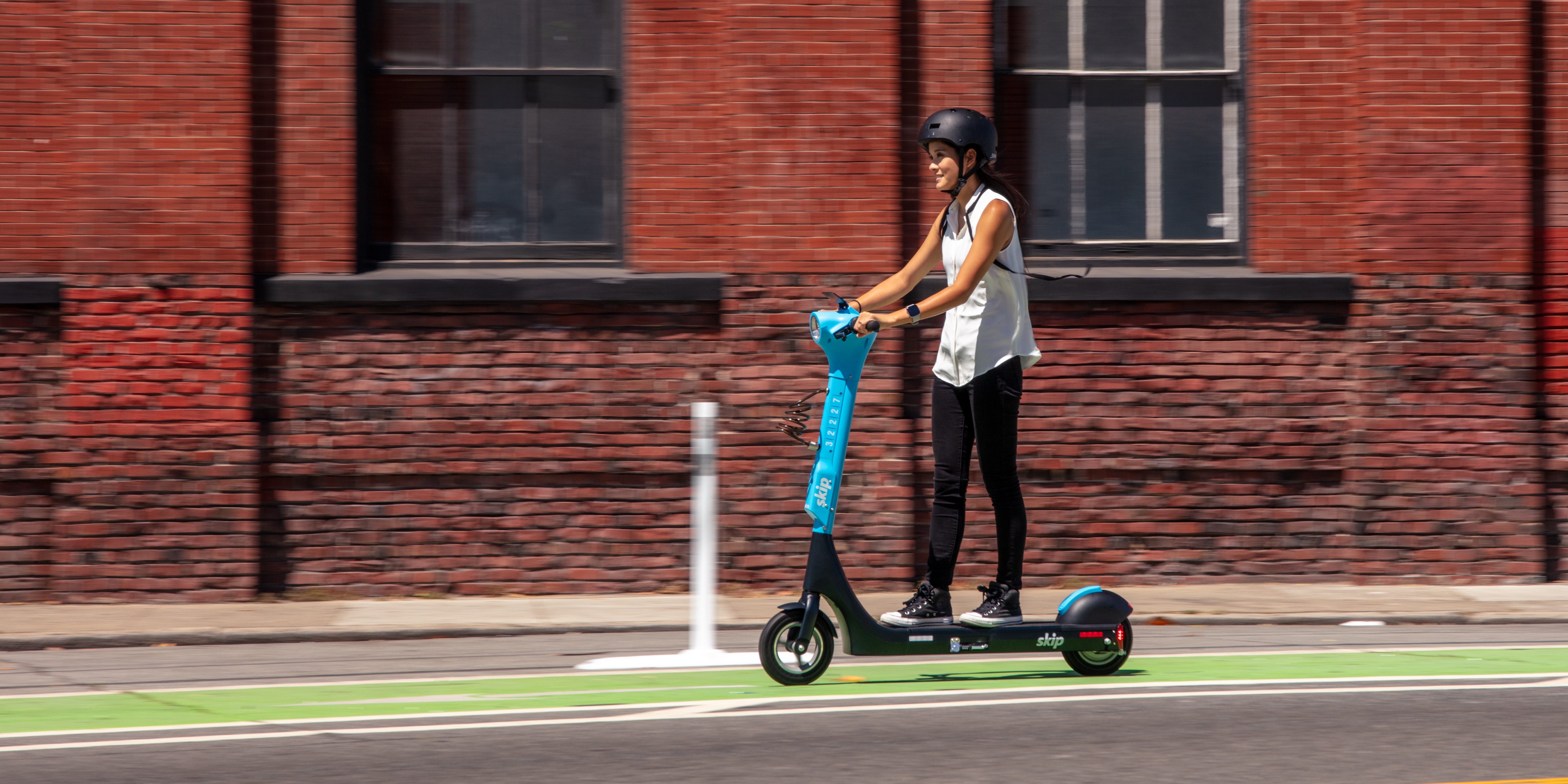 A helmeted rider on a blue Skip scooter in a green bike lane beside a brick building