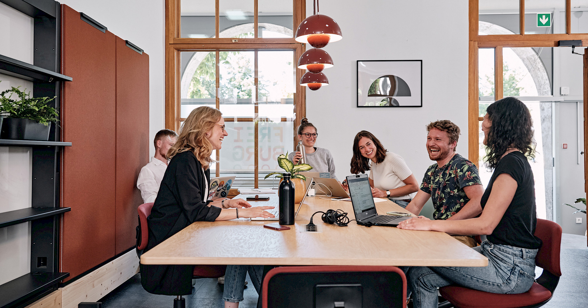 People with laptops gathered around a wooden table in a bright shared workspace