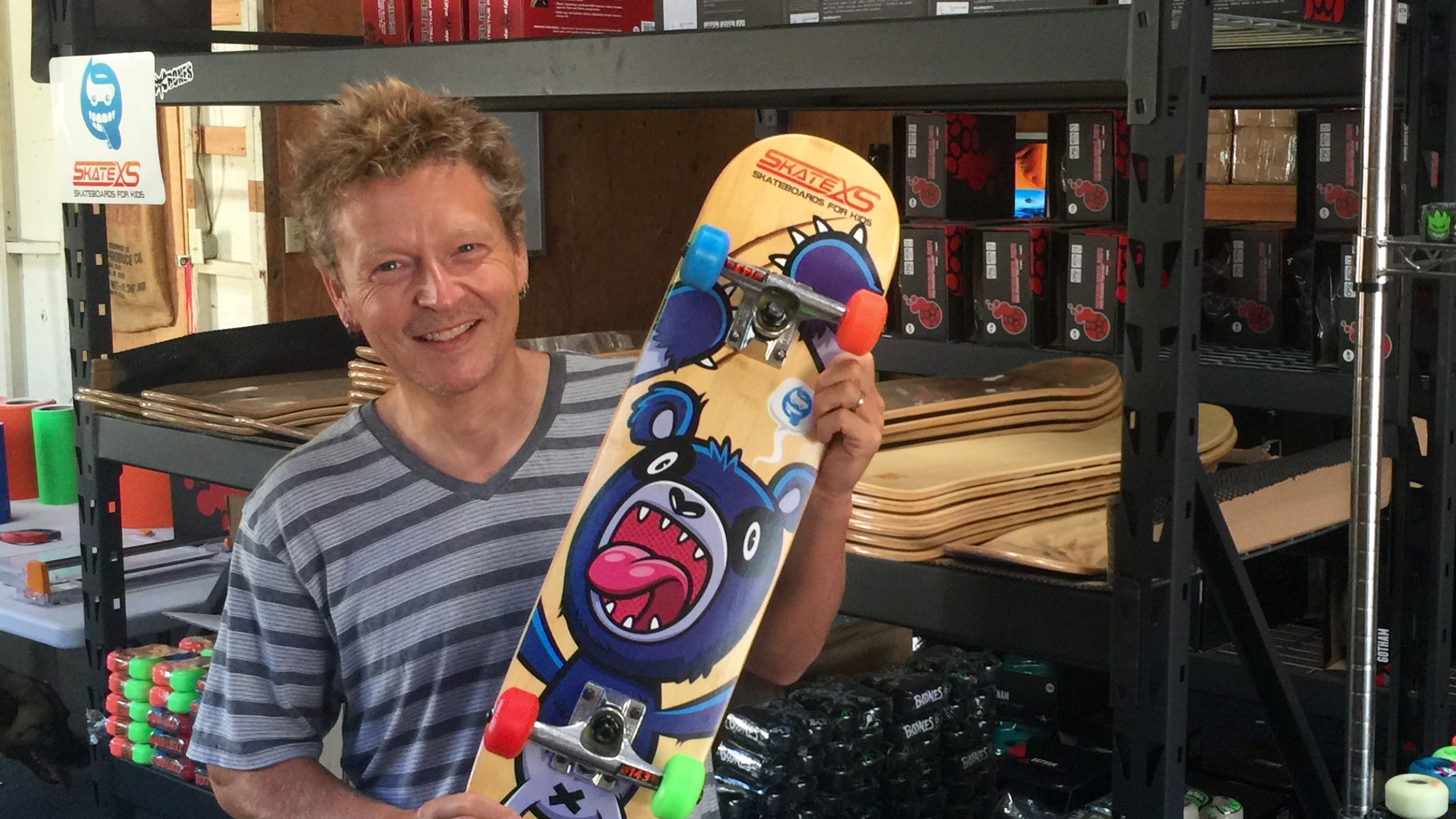 A Skate XS business owner holds a brightly illustrated children's skateboard in front of stocked shelves
